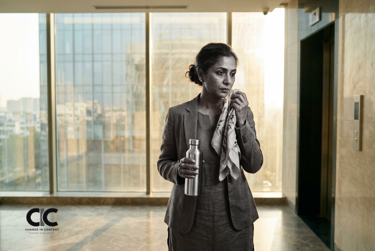 A midlife Indian woman standing in a corridor with a water bottle and scarf, representing menopause symptoms at work.