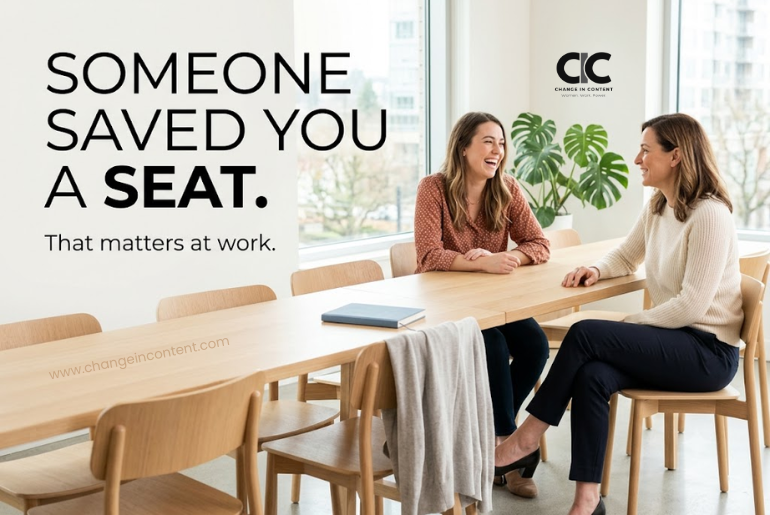 Two women colleagues in relaxed conversation at a bright workplace table, with an empty chair saved beside them, representing friendship, belonging and support at work.