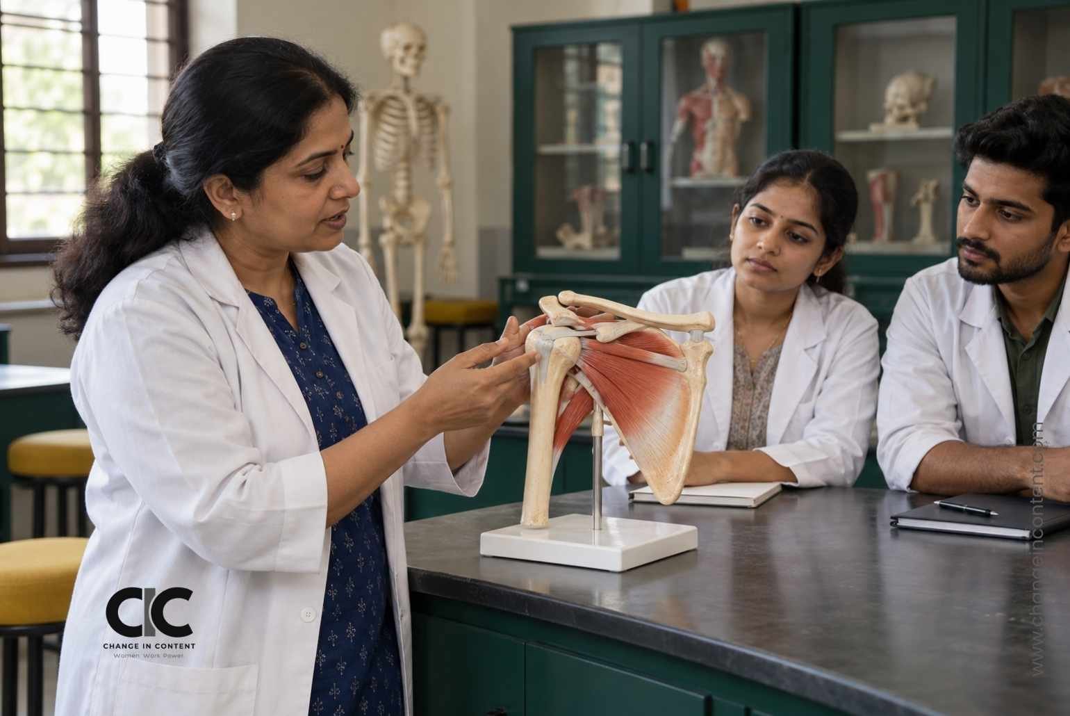 Illustrative image of a woman educator demonstrating an anatomical shoulder model to adult trainees in a medical teaching room.
